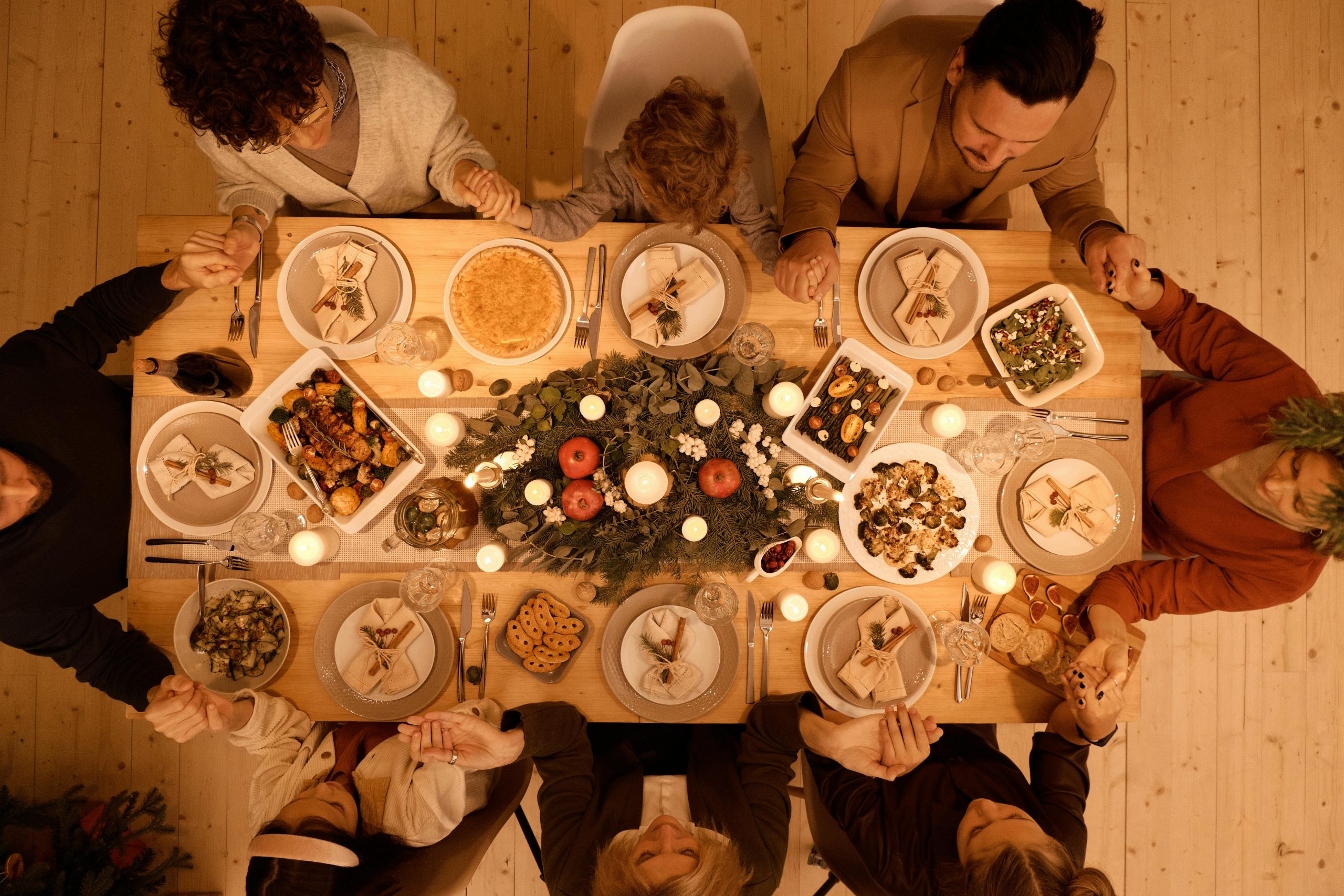 A top-down view of a dinner table with people sitting around it holding hands. There are holiday lights on the table